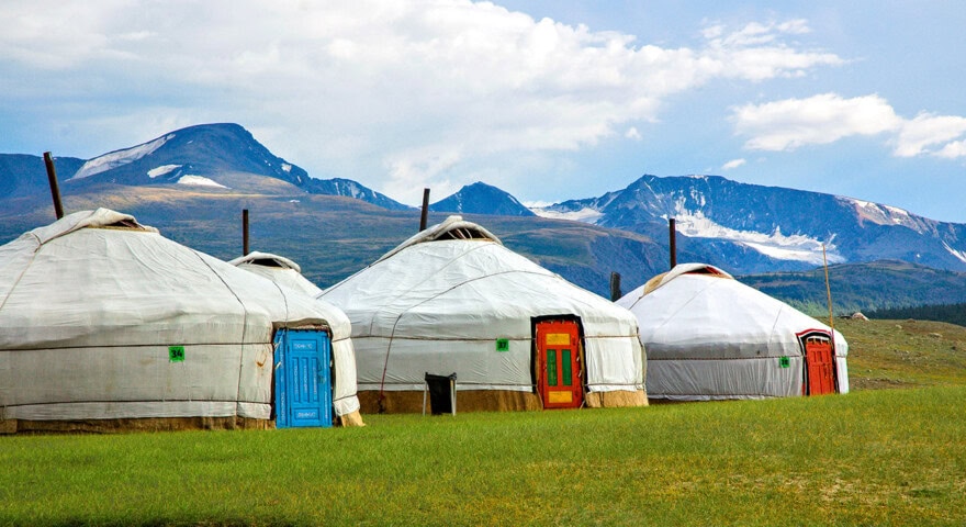 Four traditional Mongolian yurts with colorful doors are set against a backdrop of green grass and distant snow-capped mountains under a partly cloudy sky.