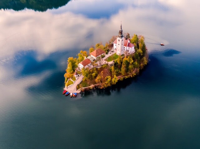 Aerial view of a small Slovenian island with a church and other buildings surrounded by a calm lake; a few boats are docked at the island, and the surrounding area is lush and green—a perfect travel destination for those passionate about tourism.