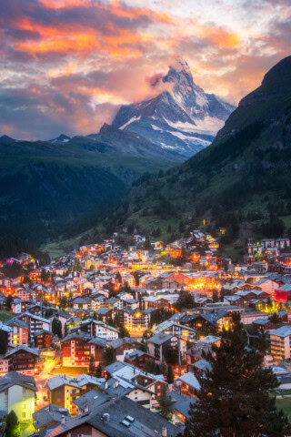 A vibrant, illuminated town is nestled in a Swiss valley with a mountain peak, partially covered in snow, under a colorful, dramatic sunset sky.