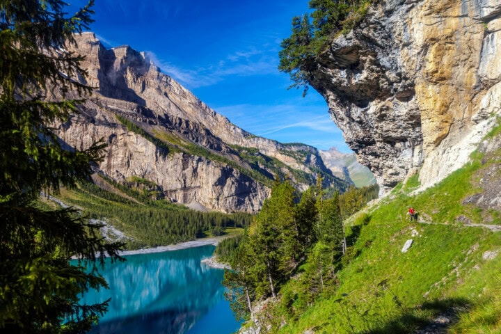 A hiker ascends a grassy hillside in Switzerland with a lake below, flanked by tall green trees and towering cliffs, under a clear blue sky, capturing the essence of travel and tourism in this picturesque landscape.