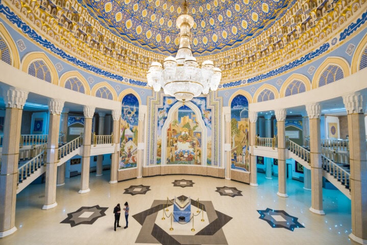 A grand hall with an ornate mosaic ceiling, a large chandelier, and intricate wall murals reminiscent of Uzbekistan's rich heritage. Two people stand near a central decorative piece surrounded by barriers.