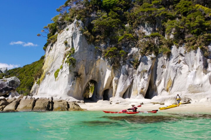 Two people kayak near a rocky shore with white cliffs and dense greenery in the background under a clear blue sky, showcasing the natural beauty of New Zealand.