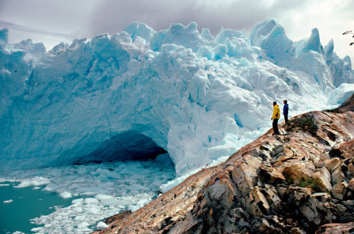 Two people wearing yellow and blue jackets stand on a rocky slope near an immense ice formation with blue hues and a cave-like opening, surrounded by icy water and Argentina’s cloud-filled sky.