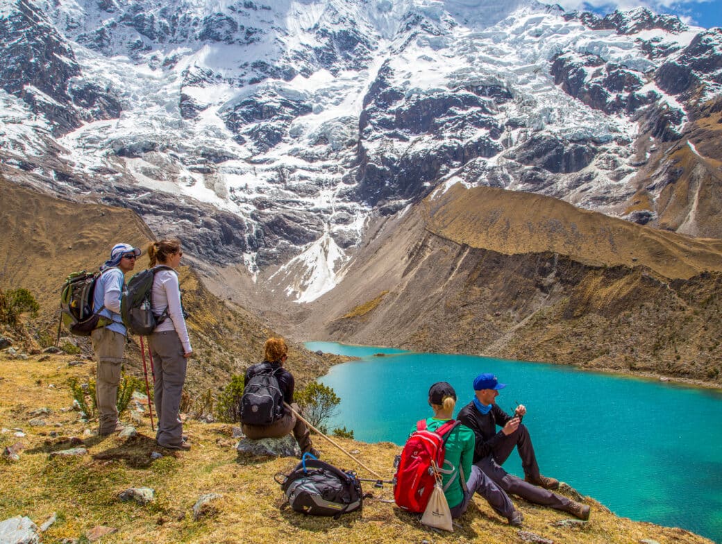Four hikers with backpacks gather near a turquoise alpine lake, surrounded by rugged snow-capped mountains. Three are standing and one is sitting on the ground, ready to embrace the breathtaking beauty of this landscape in all months.
