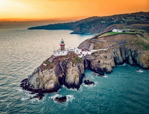A lighthouse sits atop rugged cliffs on an ocean coastline at sunset in Ireland, with surrounding hills and houses in the background.