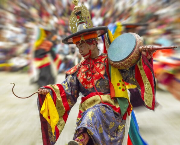 A person in a colorful traditional outfit performs a cultural dance holding a drum and a whip, with a blurred crowd of tourists in the background, capturing the spirit of travel to Bhutan.