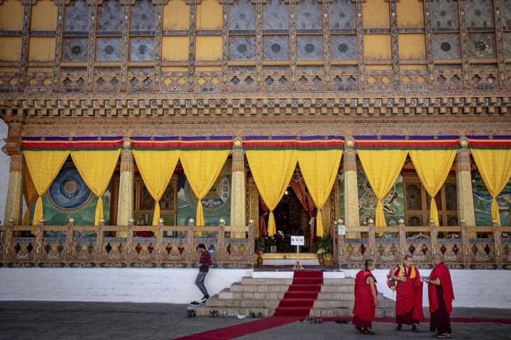 Three monks converse in front of an intricately decorated Bhutanese temple facade with yellow curtains and detailed carvings. A man sits on the steps nearby, while a red carpet leads to the temple entrance.