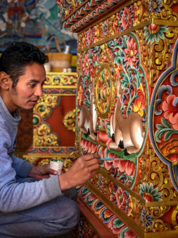 A person is meticulously painting a detailed and colorful design on a wall, featuring intricate floral patterns and animals, reflecting the rich cultural heritage of Bhutan. This artwork adds to the country's allure as a top travel destination for those seeking unique tourism experiences.