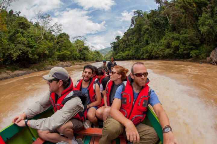 A group of people wearing life jackets are seated in a small boat traveling through a river surrounded by the dense vegetation of Malaysia.