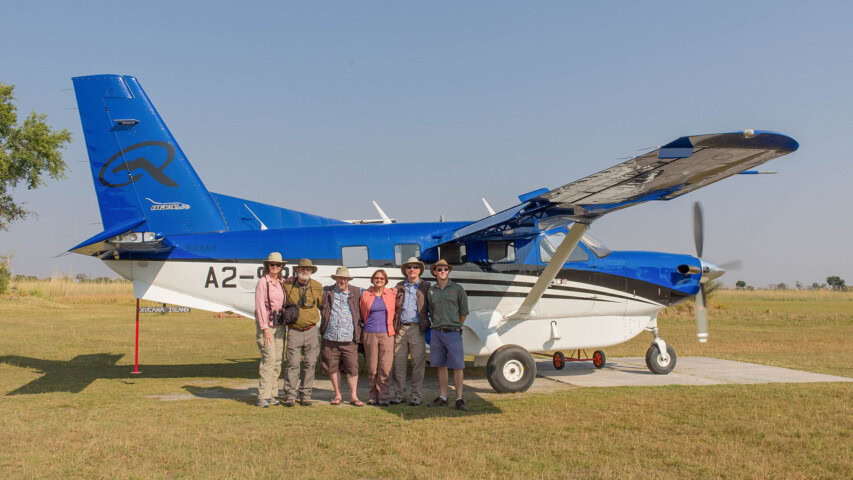 Six people dressed in outdoor attire stand in front of a small blue and white aircraft on a clear day, ready for their adventure, with grassy fields in the background.