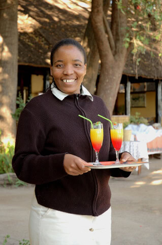 A smiling guide holding a tray with two colorful drinks garnished with straws, standing outdoors with trees and buildings in the background.