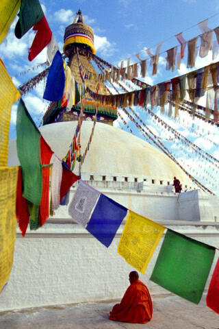 A monk in orange robes sits near the large white dome of Boudhanath Stupa in Nepal, adorned with colorful prayer flags against a blue sky in Kathmandu.