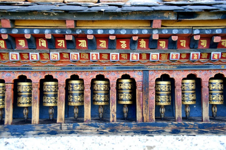 A row of Tibetan Buddhist prayer wheels featuring intricate carvings and inscriptions, set beneath a colorful wooden structure, offers a glimpse into the rich cultural tapestry that makes Bhutan an alluring travel destination.