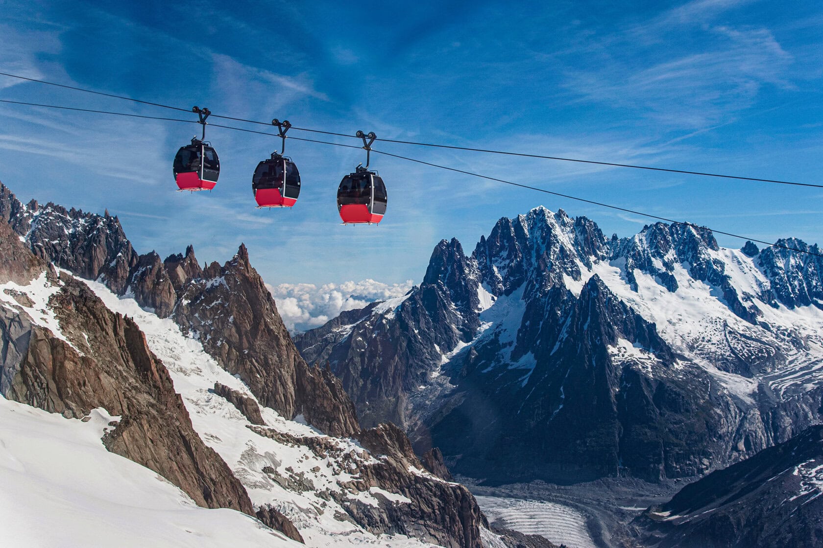Three red cable cars travel over snowy mountains under a blue sky, offering a breathtaking view for any trip, with jagged peaks and a glacier visible in the background.