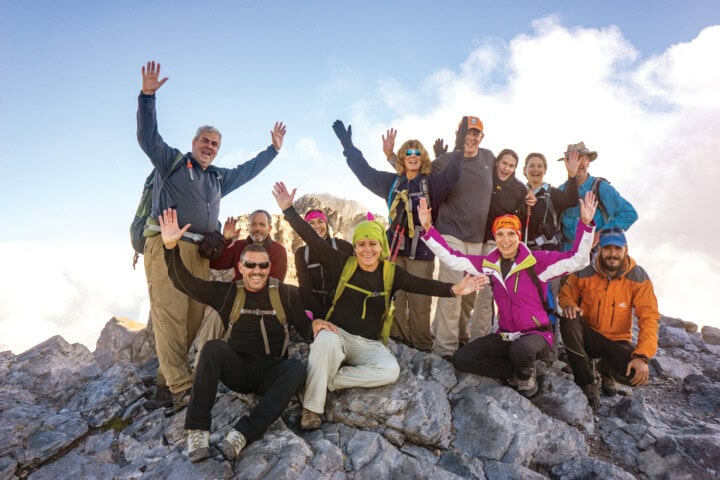A group of hikers standing on a rocky mountain summit in Greece, their arms raised and smiling at the camera, capturing the joy of travel and adventure.