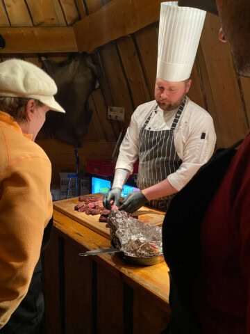 A chef in a white hat and striped apron slices meat on a wooden counter, while three tourists, captivated by the culinary art, watch closely. The scene brings to life the rich flavors of Finland's thriving food scene.