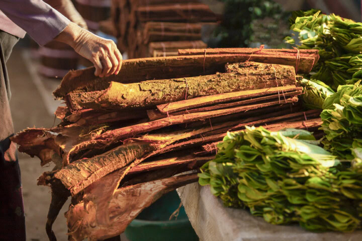 A person’s hand reaches for a pile of stacked raw cinnamon sticks next to bundles of green leaves at an outdoor market stall in Sri Lanka.