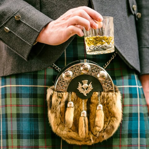A person in traditional Scottish attire, including a kilt and sporran, holds a glass of whisky, capturing the essence of Scotland and inviting you to explore its rich culture.