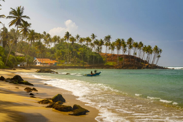 A sandy beach in Sri Lanka with rocks in the foreground, coconut trees along the shoreline, houses with red roofs, and people in a small boat on the calm sea.