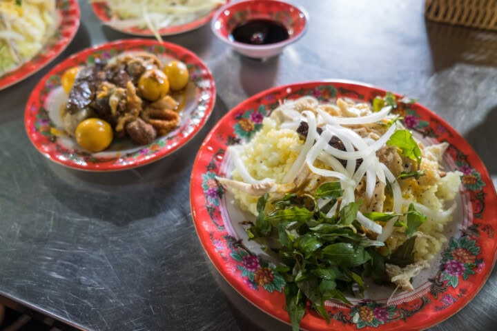 A close-up of two dishes on a metal table: a plate with rice, shredded chicken, onions, and herbs in the foreground, reminiscent of traditional Vietnamese flavors, and another plate with assorted meats and vegetables beside a small bowl of sauce in the background.
