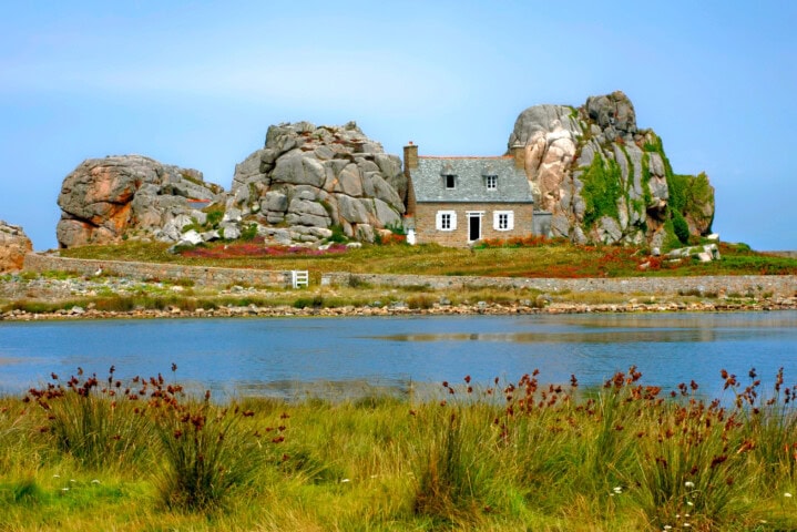 A small house with a grey roof is nestled between large boulders, situated by a body of water with grassy foreground under a clear blue sky, reminiscent of the charming countryside in France.