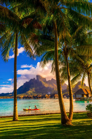Two people kayaking on clear blue water with overwater bungalows and a mountain in the background, framed by tall palm trees, capture the breathtaking essence of French Polynesia.