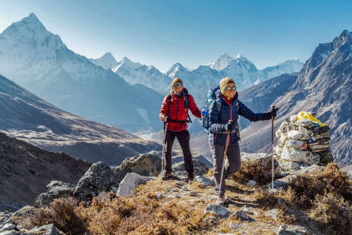 Two hikers with backpacks and hiking poles traverse a trail in Nepal, surrounded by snow-capped peaks. The clear sky and sunlight illuminate the breathtaking mountainous landscape.