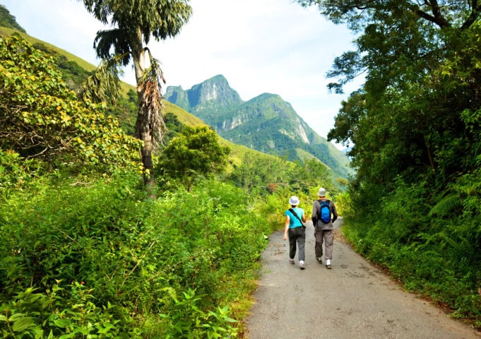 Two people with hats and backpacks walk on a path surrounded by lush greenery, with the breathtaking mountains of Sri Lanka in the distance.