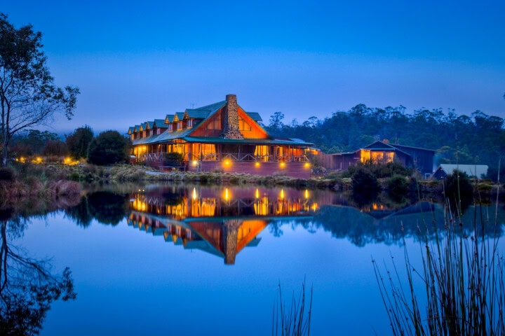 A large lodge with warm lights reflected on a still lake at twilight, surrounded by trees and bushes in picturesque Tasmania.