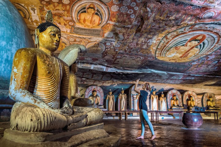 A person stands in a cave temple in Sri Lanka, decorated with numerous Buddha statues and intricate ceiling paintings, holding a phone to take a picture.