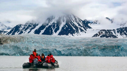 A group of people in red jackets sit on an inflatable boat on a body of water with a mountainous, snow-covered landscape and glacier in the background. This Svalbard wildlife adventure showcases the stunning beauty of the polar region.