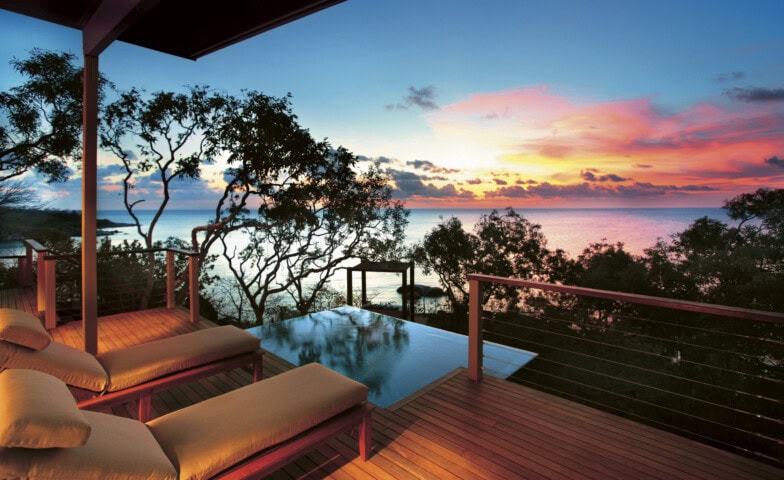 Sunset view from a wooden deck in Australia with lounge chairs overlooking an infinity pool and the ocean, surrounded by trees.
