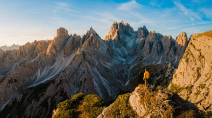 A solo traveler in a yellow jacket stands on a rocky hill, overlooking a rugged mountain range under the clear blue sky—an inspiring scene for those who seek unforgettable travel experiences.