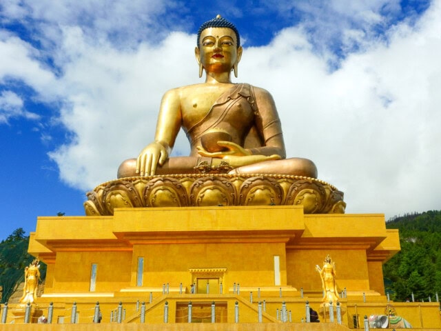 Golden statue of a seated Buddha on a large lotus pedestal against a blue sky with clouds, resembling the serene architecture found in Bhutan, surrounded by smaller statues and a railing at the base.