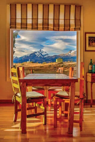 A wooden dining table with colorful chairs is positioned by a large window, framing a scenic view of Argentina's snow-capped mountains and partly cloudy sky.
