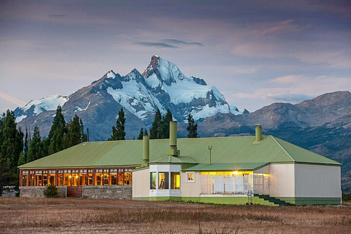 A green-roofed lodge is situated in front of a snow-capped mountain with a blend of trees and open field in the foreground, during a picturesque dusk in Argentina.