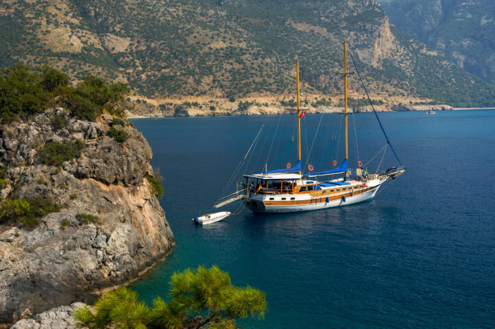 A boat is anchored near a rocky coastline with cliffs and greenery in Turkey, surrounded by clear blue water. A small dinghy floats beside the boat.