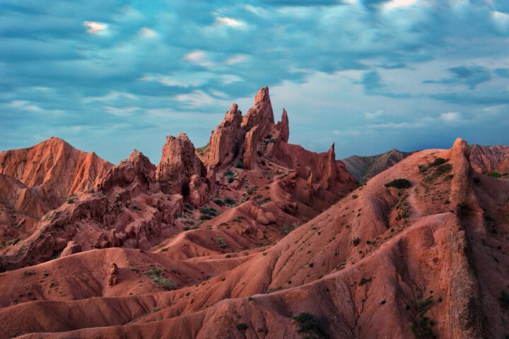 Jagged red rock formations rise sharply against a backdrop of a cloudy sky and the rugged landscape of Kyrgyzstan.