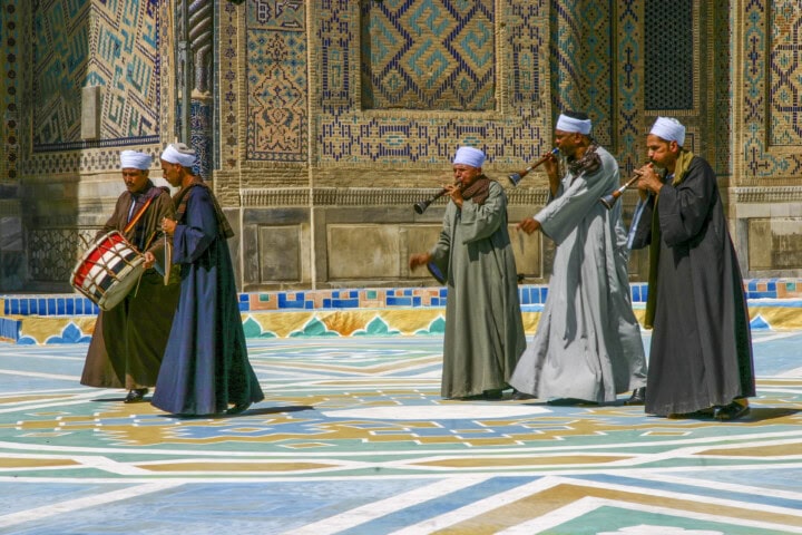 A group of five men in traditional attire perform music with wind instruments and a drum in a tiled courtyard adorned with intricate mosaic patterns, evoking the rich cultural heritage of Uzbekistan.