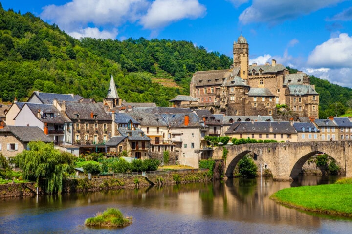 Scenic view of a historic French village with stone buildings and a castle on a hill. A stone bridge with multiple arches spans a river in the foreground. Lush green vegetation surrounds the area.