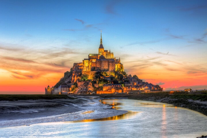 A historic castle sits atop Mont Saint-Michel, an island commune in France’s Normandy region, highlighted by sunset hues and bordered by a reflective waterway.