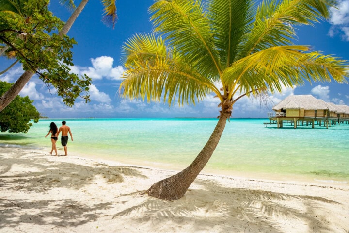 A couple walks along a tropical beach in French Polynesia, with clear turquoise water, palm trees, and overwater bungalows in the background under a sunny blue sky.