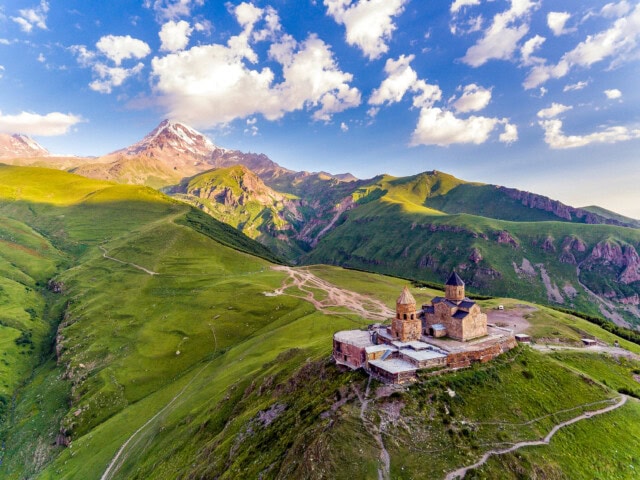 A small, old church is situated on a grassy hilltop with winding paths in the charming state of Georgia, surrounded by expansive green mountains and a partly cloudy sky.