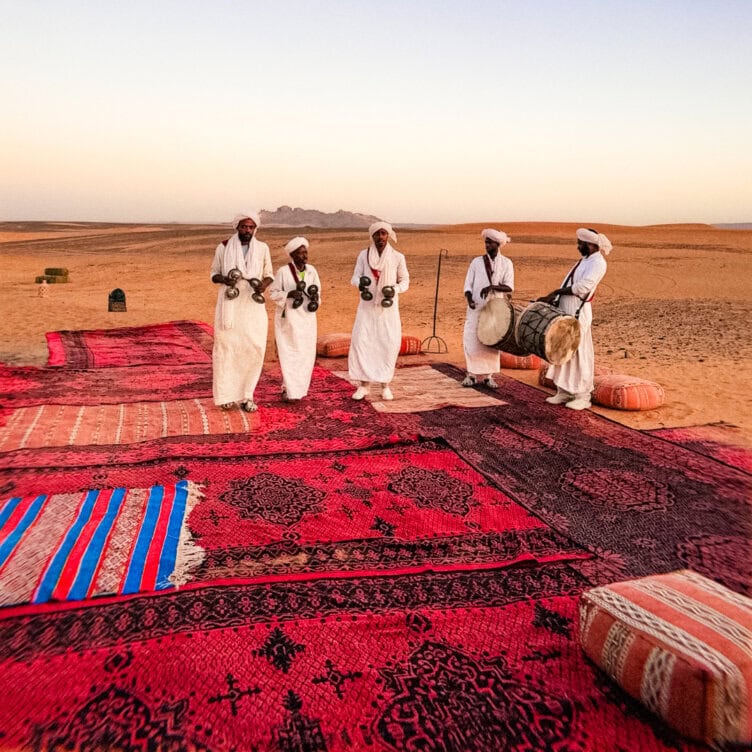 Five men in white robes play traditional instruments on patterned rugs in a desert at sunset, creating not to miss moments against distant mountains—an unforgettable scene awaiting you in 2026.