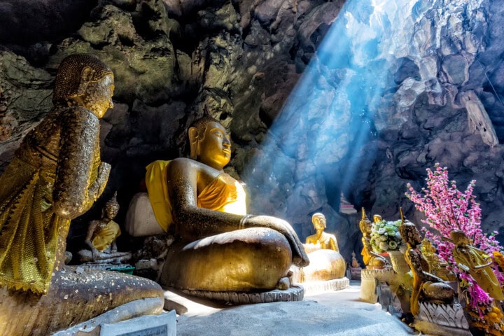 Golden Buddha statues seated inside a sunlit cave, with rays of light streaming down from above, illuminating the figures. Pink flowers are visible on the right side of the image, evoking a serene atmosphere reminiscent of sacred sites in Sri Lanka.