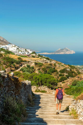 A person with a purple backpack walks down stone steps towards a picturesque Greek coastal village with white buildings and greenery, overlooking the azure sea and distant island.