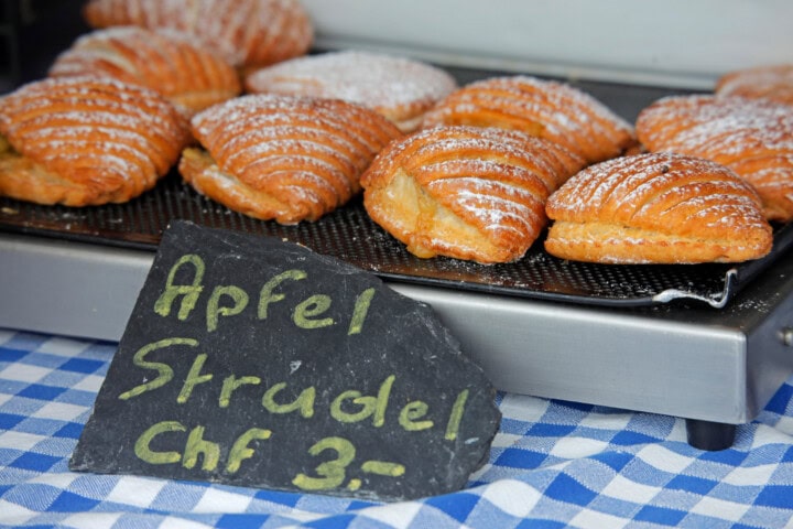 Pastries labeled "Apfel Strudel" priced at 3 CHF each are displayed on a tray with a blue and white checkered cloth underneath, reminiscent of charming Switzerland.
