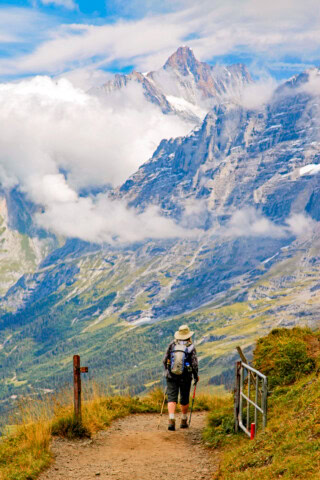 A person with hiking gear walks down a mountain trail in Switzerland, surrounded by tall, snow-capped peaks and a valley covered in clouds—a perfect scene for tourism enthusiasts craving adventure.