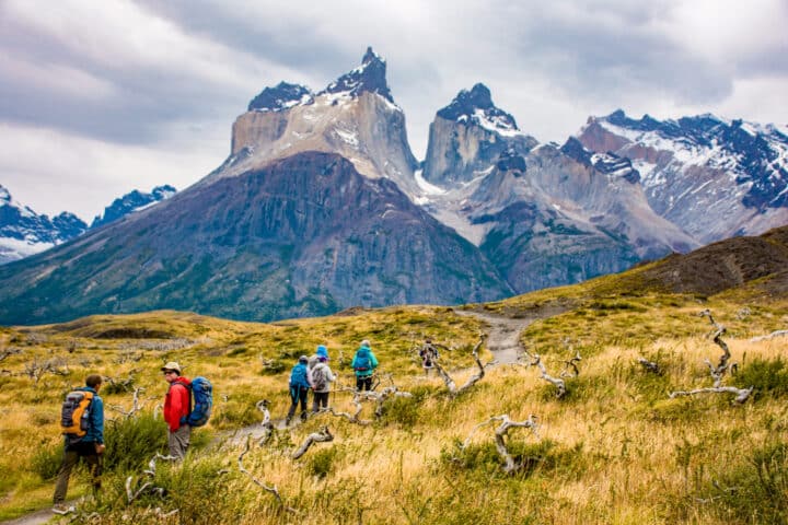 Hikers with backpacks walk on a trail through the grassy terrain of Chile, with mountains in the background under a cloudy sky.