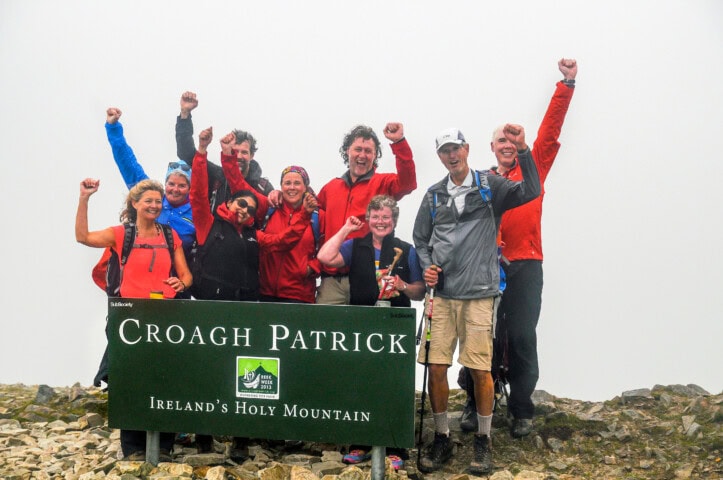 A group of hikers stands triumphantly at the summit of Croagh Patrick mountain, raising their arms in celebration. A sign in front reads "Croagh Patrick, Ireland's Holy Mountain," capturing the spirit of travel and tourism in this beautiful part of Ireland.